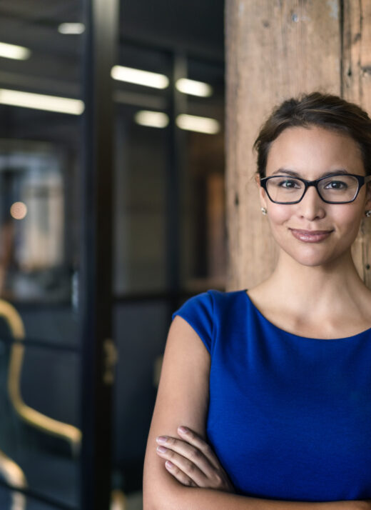 Portrait of confident businesswoman with arms crossed in office. Female professional is smiling at workplace. Beautiful executive wearing eyeglasses is in formals.