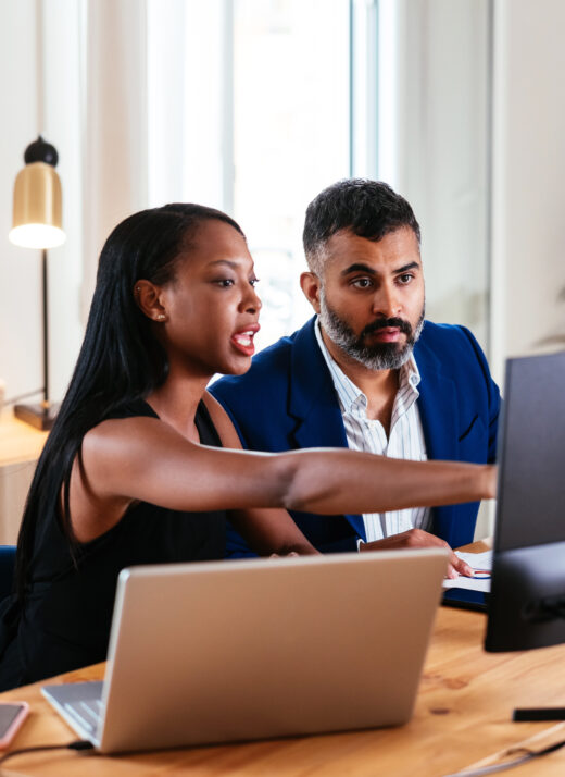 Smart black businesswoman pointing on computer screen while sharing project details with mature serious bearded colleague in suit. They are sitting at table in office room and discussing work.